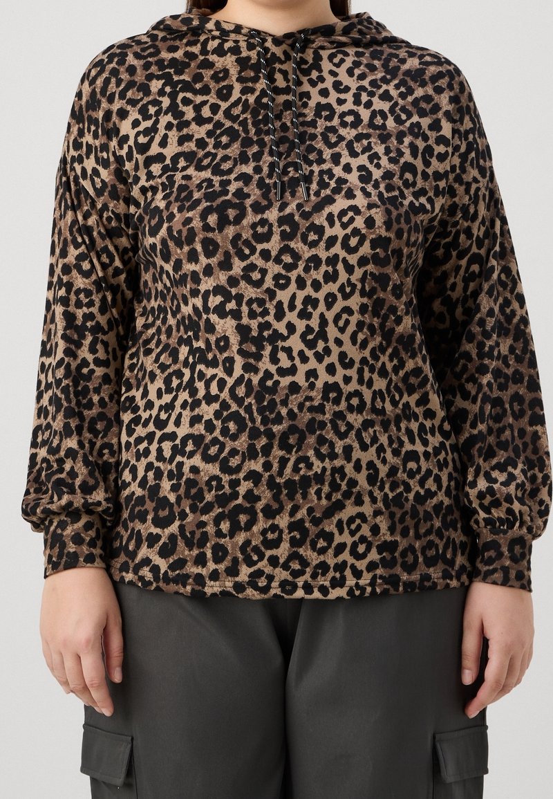 Personne portant un sweat à capuche imprimé léopard à manches longues avec cordons et un pantalon cargo sombre, debout devant un fond uni.