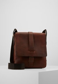 Hnědá kožená taška přes rameno s předním klopem, nastavitelným látkovým popruhem přes rameno, vyraženým logem a minimalistickým designem.
