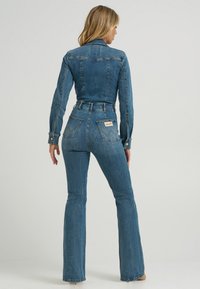Denim jumpsuit i mellomblå med en tettsittende kropp, lange ermer og vide bukseben. Har påsydde lommer og kontrastsømmer som detaljer.