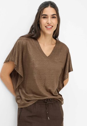 Ung kvinde smilende, iført en glitrende brun V-hals top med korte flæseærmer og mørkebrune bukser med snøre i taljen.