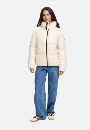 Cream puffer jacket with high collar, front zip, and black accents. Model wears blue jeans and sneakers. Smooth, quilted texture.