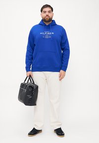Man in een blauwe Tommy Hilfiger hoodie, witte broek, zwarte sneakers, met een zwarte, gestructureerde handtas met dubbele handvatten.
