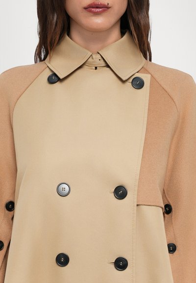 Beige double-breasted coat with a collared neckline, featuring contrasting tan fabric on shoulders and black buttons. Smooth texture.