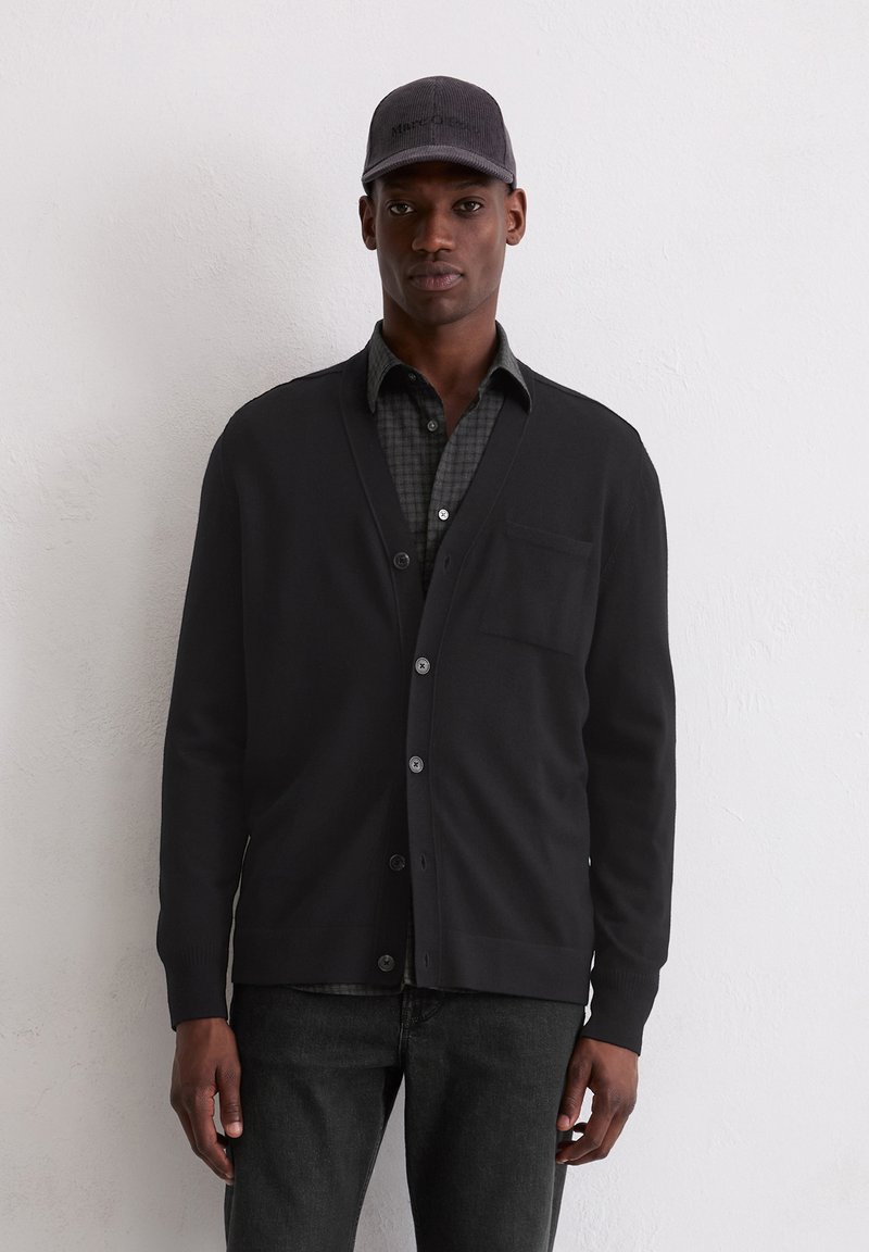 Schwarzer Cardigan mit Knöpfen, hergestellt aus weichem Material, mit V-Ausschnitt und einer Fronttasche. Getragen über einem karierten Hemd und schwarzen Jeans.