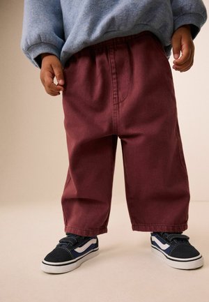 Kleinkind trägt lockere bordeauxrote Hose, blau-grauen Pullover und schwarz-weiße Sneaker und steht vor hellem Hintergrund.