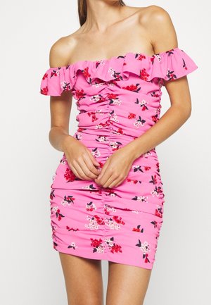 Femme portant une mini-robe rose froncée, à épaules dénudées, avec des imprimés floraux rouges et blancs, mains jointes à la taille.