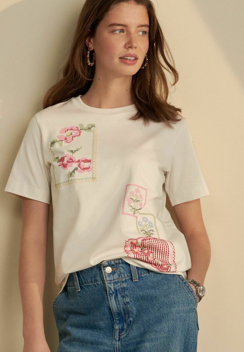 T-shirt en coton blanc avec broderies florales en rose et vert, présentant divers motifs et accents sur le devant. Manches courtes, col rond.