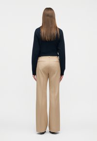 Άτομο με μακριά ίσια μαλλιά, που φοράει μαύρο πουλόβερ, μπεζ παντελόνια με φαρδιά πόδια και μαύρα τακούνια, στέκεται με την πλάτη προς τα πίσω πάνω σε λευκό φόντο.