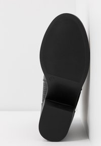 Semelle d'une botte noire avec une bande de roulement texturée et un talon bloc, présentée à la verticale sur un fond blanc d’angle.