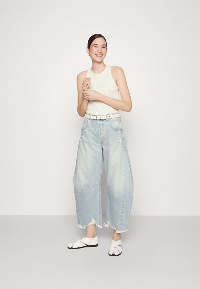 Pantaloni in denim blu chiaro a gamba larga con orli frangiati e dettagli con effetto usurato, abbinati a un top senza maniche bianco e scarpe bianche con cinturini incrociati.