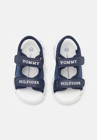 Námorně modré sandály pro batolata s bílými podrážkami, vyrobené z textilu a syntetického materiálu. Obsahují dva nastavitelné suché zipy se značkovými logy.