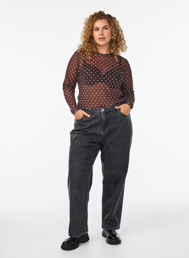 Vrouw met krullend blond haar die een doorschijnend zwart polkadot topje draagt over een zwarte bh, losse zwarte spijkerbroek en zwarte schoenen, staand met handen in de zakken.
