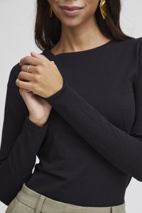 Kvinna klädd i svart långärmad topp och olivgröna byxor, med händerna sammanlänkade nära bröstet, och synliga guldfärgade örhängen formade som blad.