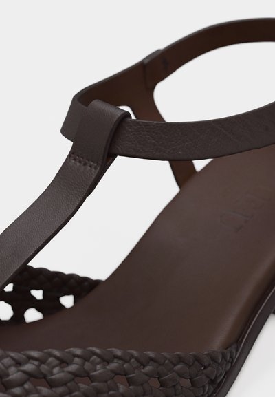 Καφέ δερμάτινο σανδάλι με λουράκι σε σχήμα Τ και πλεκτή λεπτομέρεια στο μπροστινό μέρος πάνω σε λευκό φόντο.