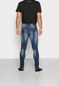 Jeans blu in denim slim-fit con dettagli strappati e accenti di vernice splatter, abbinati a una semplice t-shirt nera e scarpe da ginnastica nere.