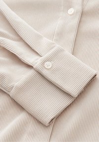 Tissu de chemise ribbed beige avec un poignet boutonné et une texture subtile. Présente un bouton standard de couleur assortie. Design épuré et simple.