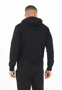 Sudadera negra con capucha, de mangas largas, con puños y dobladillo de canalé. Textura suave con un bolsillo tipo canguro y cordón ajustable. Vista trasera.