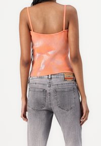 Femme portant un haut sans manches orange brillant et un jean gris clair avec un patch en cuir de la marque Diesel à l'arrière, côté droit de la ceinture.