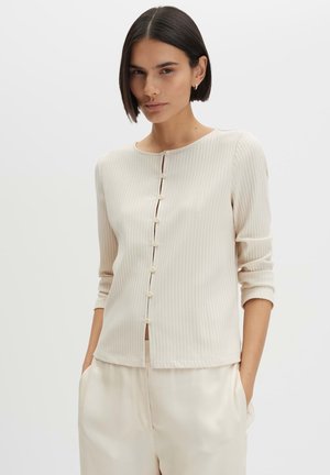 Vrouw met kort donker haar, draagt een crèmekleurig geribd gebuttond vest en crèmekleurige losse broek, met handen in de zakken, tegen een effen achtergrond.