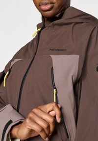 Καφέ μπουφάν με γκρι λεπτομέρειες, κατασκευασμένο από ελαφρύ υλικό. Διαθέτει φερμουάρ μπροστά μ ε κίτρινα εξαρτήματα και κουκούλα. Κοντινό πλάνο χεριού που ρυθμίζει το φερμουάρ.