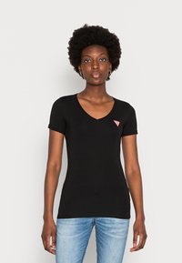 T-shirt nera a maniche corte con scollatura a V e design aderente. Presenta un piccolo logo triangolare rosso sul petto sinistro. Abbinato a jeans azzurri chiari.