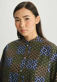 Chemise à boutons présentant un mélange de motifs floraux et géométriques dans des teintes de vert foncé, bleu et marron. Tissu doux avec un col et des boutons.