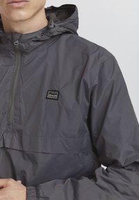 Gray hooded jacket made of lightweight, smooth fabric. Features a front zipper and a small logo patch on the chest.