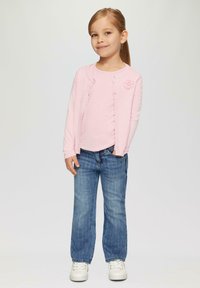 Helle rosa Strickjacke mit floralem Akzent, kombiniert mit einem hellrosa Top und blauen Denim-Jeans. Ausgestattet mit weißen Sneakers und einem schlichten Design.