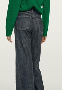 Jeans a gamba larga in denim scuro con vita alta, caratterizzati da cuciture rosa a contrasto e due tasche posteriori, indossati con un maglione verde.