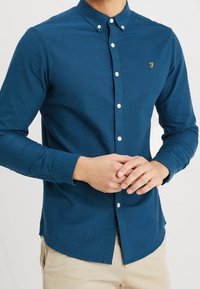 Koszula z długim rękawem, zapinana na guziki, w ciemnoniebieskim kolorze, wykonana z bawełny o dopasowanym kroju. Ma białe guziki oraz małe złote logo na klatce piersiowej.