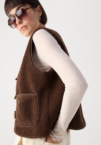 Brown fleece vest with a textured surface, featuring a chest pocket and large buttons, worn over a ribbed long-sleeve top.