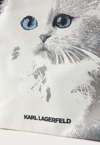 Biała torba tote z srebrnym wzorem kota i dużymi niebieskimi oczami. Nazwa marki "KARL LAGERFELD" jest wydrukowana na czarno na dole.