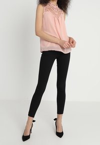 Top sin mangas de color rosa claro con detalles de encaje en el escote y un dobladillo con forma de ondas, combinado con pantalones negros ajustados y tacones negros.