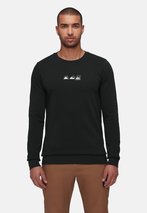 Man met een zwart shirt met lange mouwen met witte berglogo's en een bruine broek, staand tegen een effen lichte achtergrond.