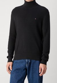 Schwarzer, strukturiert gestrickter Rollkragenpullover mit Rippenmuster und enganliegenden Bündchen. Verfügt über ein kleines Logo auf der Brust. Kombiniert mit blauen Jeans.