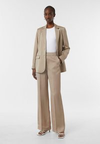 Beżowy garnitur szyty na miarę z marynarką zapinaną na jeden guzik i szerokimi spodniami. Pod spodem widoczny biały top z prążkowanej tkaniny. Dopełnieniem stylizacji są sandały na obcasie z paskami.