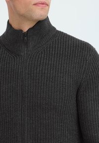 Mørkegrå strikket zip-up genser med høy krage, med et strukturert mønster og en subtil glans på stoffet.
