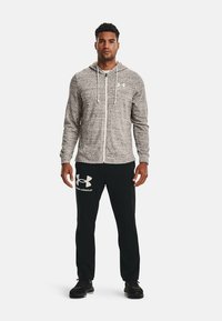 Lys grå zip-up hoodie lavet af tekstureret stof med snore. Sorte bukser med hvid "Under Armour" logo på venstre lår. Sorte sko.
