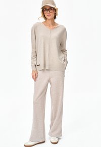 Kobieta stojąca, ubrana w beżowe spodnie o szerokich nogawkach, dopasowany luźny sweter, beżowy kapelusz typu bucket, duże okulary i białe trampki.