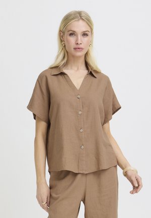 Blonde woman wearing loose brown button-up short-sleeve shirt and matching pants, standing against a plain light background.