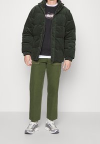 Chaqueta acolchada de color verde oscuro con un cuello alto, textura acolchada; llevada sobre un sudadera negra y pantalones verdes oliva, combinada con zapatillas grises.