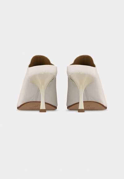 Λευκά ψηλοτάκουνα mule με μυτερά στιλέτο τακούνια και λεία υφή δέρματος, εμφανιζόμενα από πίσω σε απλό φόντο.