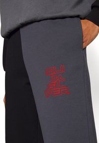 Primer plano de pantalones bicolor negro y gris con un logotipo estilizado en rojo de "Quiksilver" en la pierna gris y una mano en el bolsillo lateral.