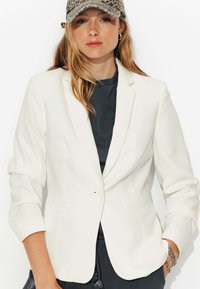 Blazer blanc ajusté avec des revers, fermeture à un bouton, et deux poches avant, porté sur un haut gris foncé.