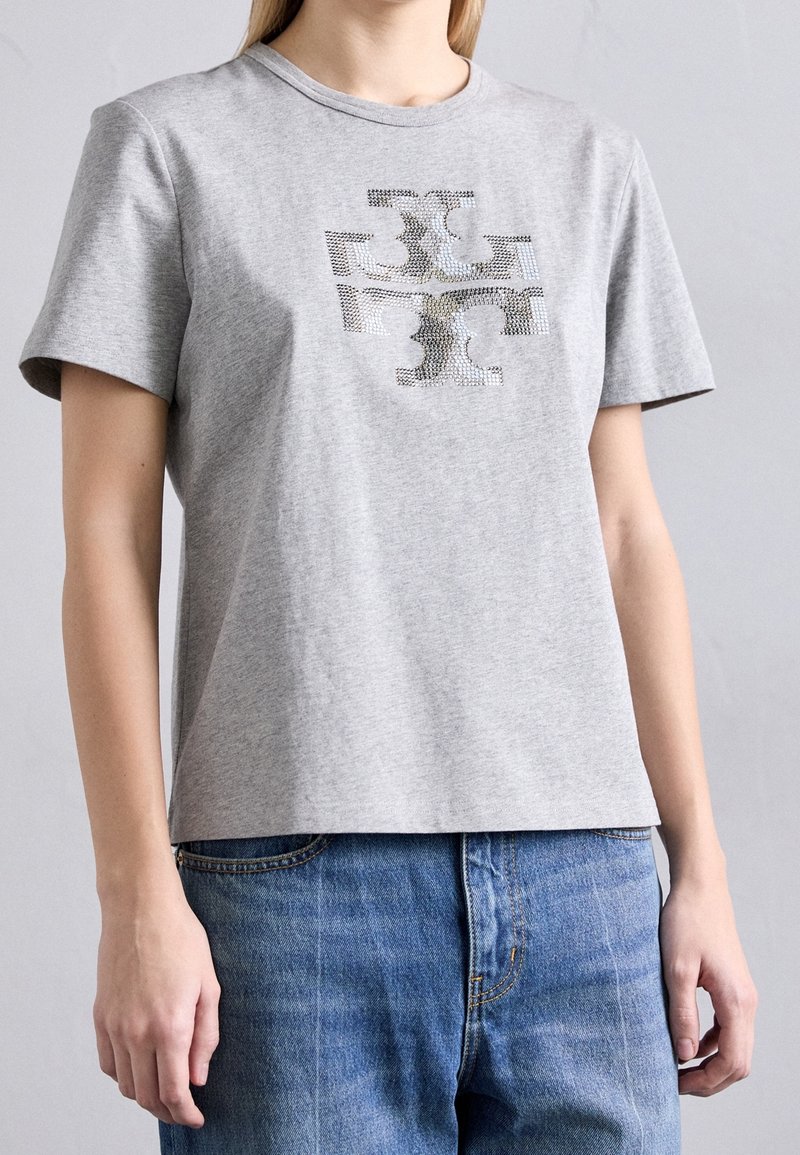 Femme portant un t-shirt gris clair à manches courtes avec un logo géométrique abstrait noir et blanc sur le devant, assorti à un jean bleu.