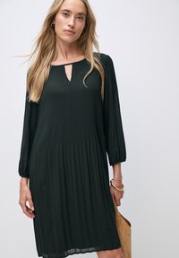 Dunkelgrünes, plissiertes Kleid mit einem Schlüsselloch-Ausschnitt, langen elastischen Ärmeln und einer fließenden Silhouette. Glatte Textur und knielanger Schnitt.