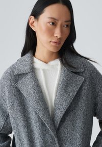 Grauer Wollmantel mit strukturiertem Finish, breiten Revers und lässiger Passform, über einem weißen, flauschigen Hoodie getragen.