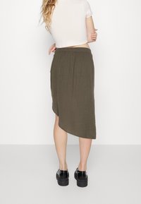 Jupe asymétrique vert olive en tissu doux avec une taille élastique, longueur genou à l'avant et plus longue à l'arrière. Chaussures à plateforme noires.