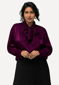 Blusa di raso viola con un grande fiocco al colletto, maniche lunghe con polsini abbottonati e una texture liscia e lucida.