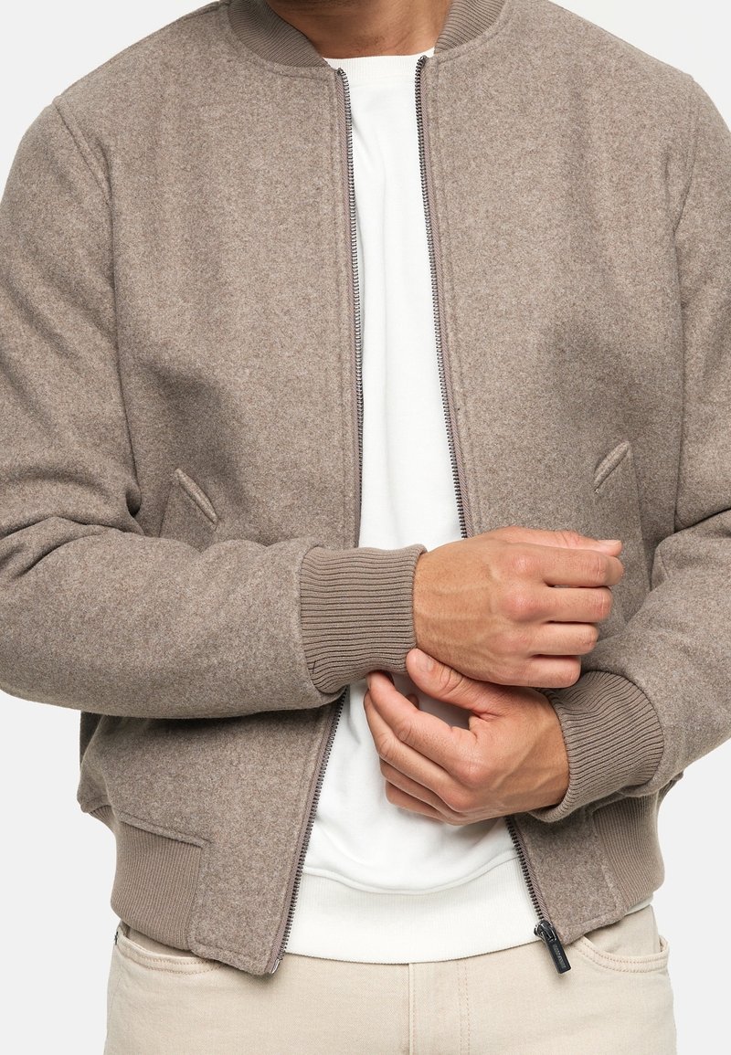Homme portant une veste en laine beige zippée sur une chemise blanche, ajustant son poignet, avec pantalon beige visible de la taille jusqu'à mi-cuisse.